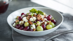 Ein Teller mit einer Portion Trauben-Feta-Salat mit Pekannüssen garniert mit ein paar Blättern frischem Basilikum. Der Teller steht auf einem grauen Küchenhandtuch, das auf einer grauen Oberfläche steht. Auf dem Tuch liegt auch noch ein Besteck. Im Hintergrund kann man noch ein Glasgefäß mit Traubensaft oder Wein zu sehen.