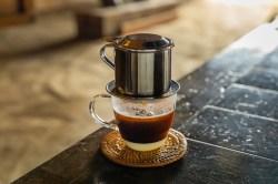 Ein Glas vietnamesischer Kaffee mit Filter steht auf einem schwarzen Holztisch. Man kann deutlich die Kondensmilch im Kaffee erkennen.